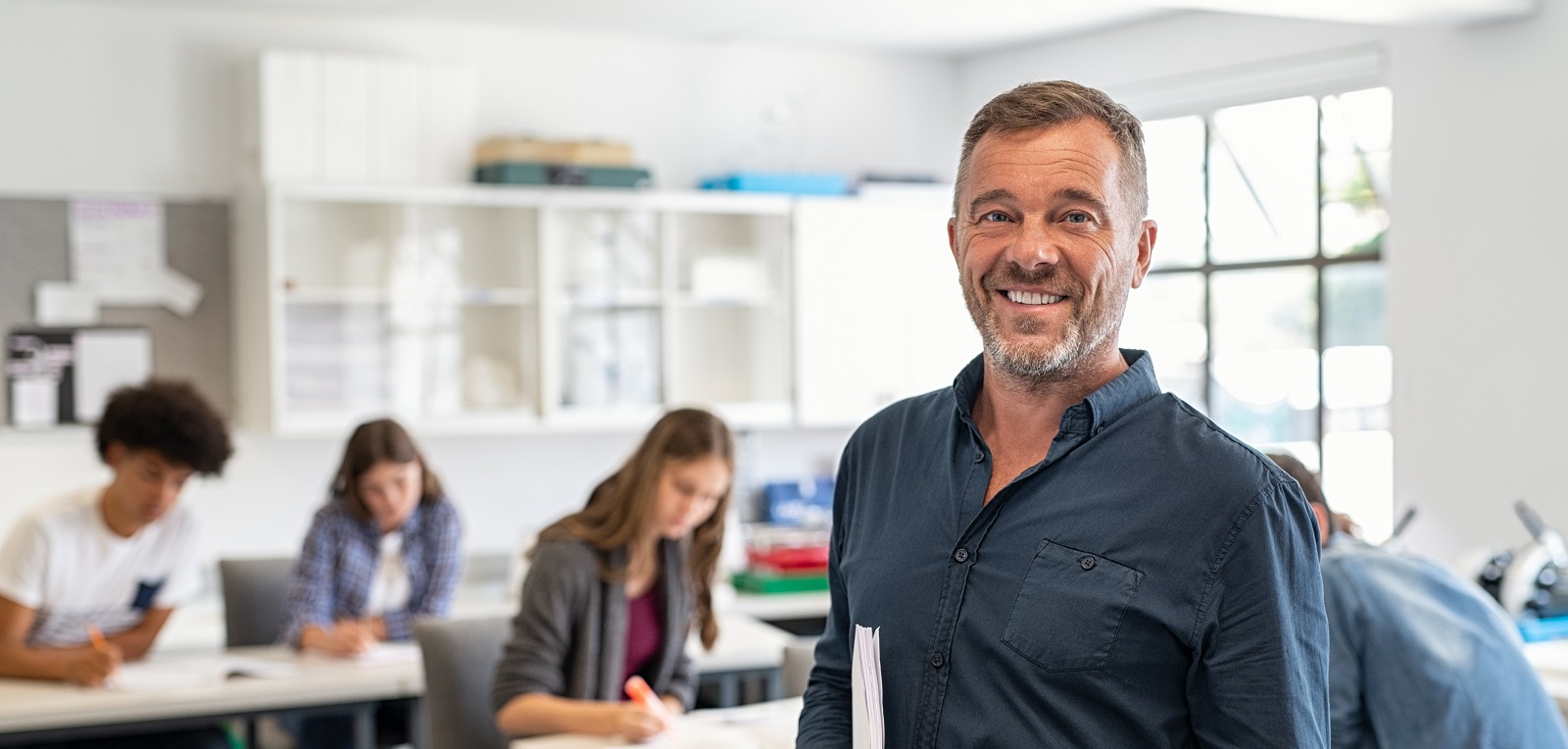 Portrait of mature teacher standing in university library and looking at camera with copy space. Happy mid adult lecturer at classroom standing after giving lecture. Satisfied high school teacher smiling and looking at camera while his studets studying in background.